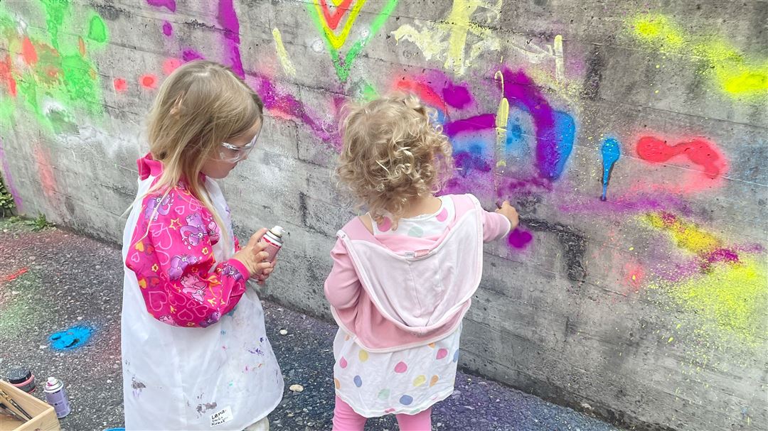 Zwei kleine Kinder mit Schutzbrille und Malschürze besprühen eine Betonwand mit bunten Sprühfarben.