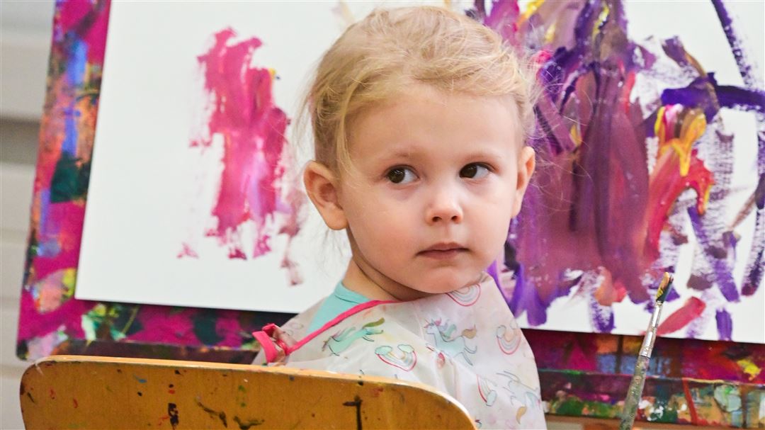 Kleinkind mit Malschürze schaut neugierig in die Kamera, umgeben von bunten Kindermalereien.