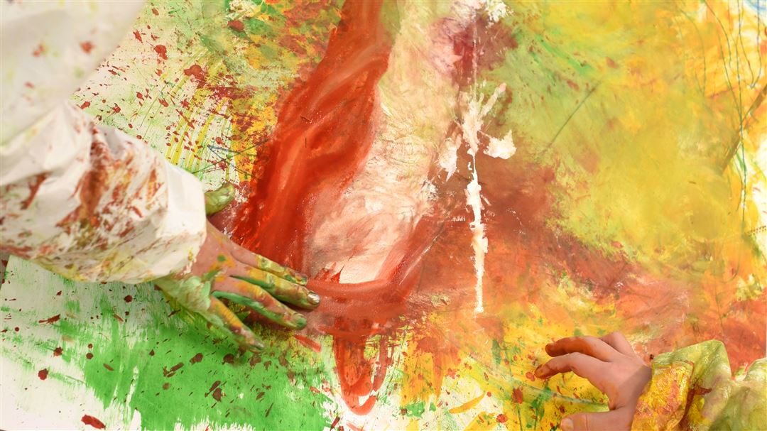 Kinderhände malen mit Fingerfarben auf einem grossformatigen Papierbogen in leuchtenden Rot-, Orange-, Gelb- und Grüntönen.