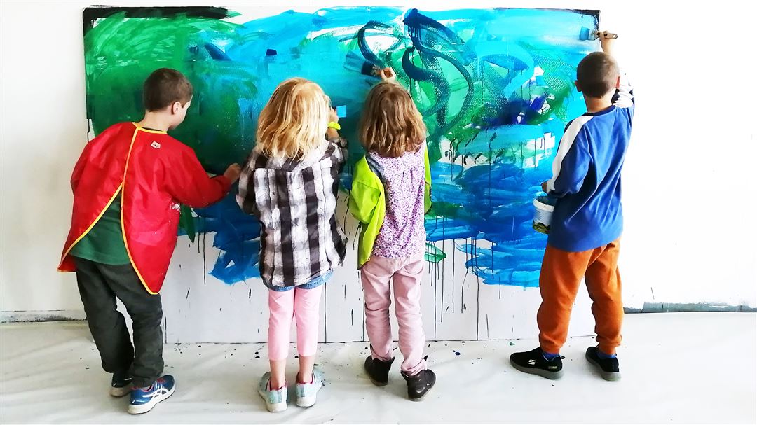 Vier Kinder malen gemeinsam mit Pinseln und Farbe an einer grossformatigen Leinwand an der Wand.